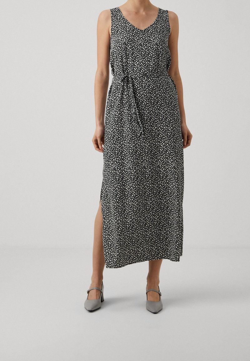 Femme portant une robe maxi sans manches noire avec un imprimé floral blanc et une fente sur le côté, accompagnée de chaussures à talons gris.