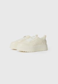 Cream-colored sneakers with a smooth leather upper, textured platform sole, and flat laces. Features a raised logo on the heel.