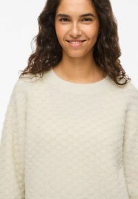 Pull tricoté couleur crème avec un motif en nid d'abeille texturé et un col arrondi. Le tissu semble doux et léger.