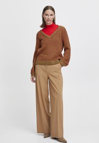 Pull en tricot marron à col en V et accents rouges, associé à un pantalon large beige et des escarpins à bout pointu. Matériaux lisses et texturés.
