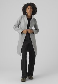 Manteau en mélange de laine gris avec un col haut et des boutons-pression, porté sur un haut noir et un pantalon noir, avec une coupe droite et des manches longues.
