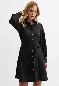Abito camicia nero realizzato in un tessuto leggero e strutturato. Presenta una chiusura frontale con bottoni, maniche lunghe, vita aderente con cintura annodata e righe verticali.