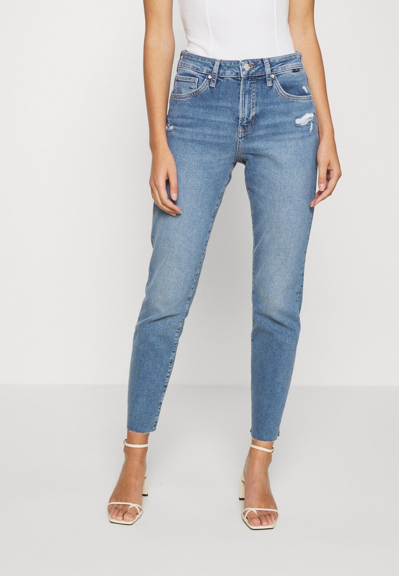 Jeans skinny de color azul claro en denim con parches desgastados y detalles destruidos. Cuenta con una cintura alta y un dobladillo sin rematar. Estilizados con sandalias blancas.