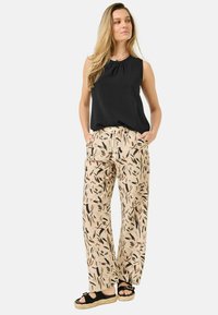Černý bezrukávový top se skládaným výstřihem, spojený s béžovými širokými kalhotami se vzorem, které mají černé a hnědé listové designy, a černými espadrilkami.