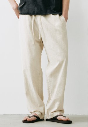 Homme portant un pantalon ample en lin beige avec un cordon, des tongs noires et une chemise foncée, les mains dans les poches, sur un fond uni.