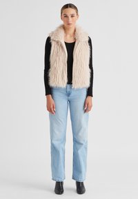 Ärmellose, hellrosa Fake-Fur-Weste mit hohem Kragen, getragen über einem schwarzen Langarmoberteil, kombiniert mit hellblauen Jeans und schwarzen Knöchelstiefeln.