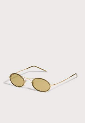 Lunettes de soleil ovales dorées, avec des verres teintés olive et une fine monture en métal, sur un fond clair.