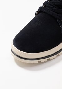 Zapato de ante azul oscuro para hombre con parte superior texturizada, entresuela color crema y suela negra robusta. Presenta cordones planos y detalles de costura.