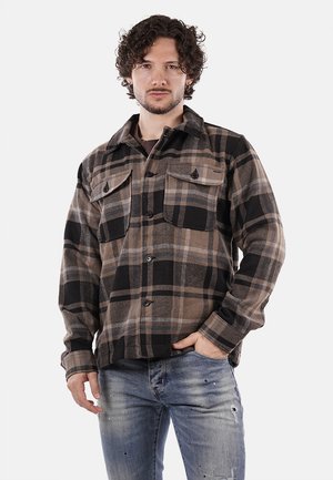 Camicia flanella a quadri nei toni del marrone, nero e beige. Presenta due tasche sul petto e chiusura con bottoni. Indossata sopra una maglietta scura con jeans strappati.