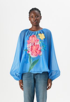 Blå skjorte i gjennomsiktig stoff med ballongsleeves som har et livlig blomstertrykk i rosa, gul og grønn. Kombinert med blå jeans.