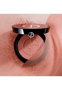 Blush compact avec un couvercle noir brillant arborant un logo. Teinte pêche douce avec une texture lisse, sur fond pêche.