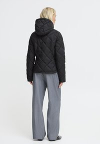 Veste matelassée noire avec capuche, arborant un motif en losanges. Associée à un pantalon large gris. Vue arrière, fond minimal.