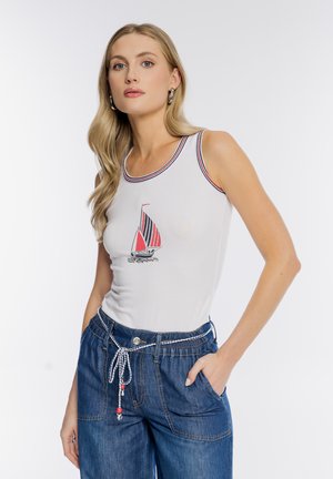 Młoda kobieta w białym topie na ramiączkach z nadrukiem czerwono-czarnej żaglówki oraz niebieskich jeansach z paskiem ze sznurka, pozująca z jedną ręką w kieszeni.