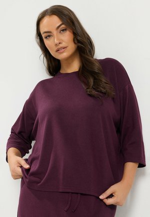 Vrouw met lang bruin haar draagt een losse donkerpaarse top en bijpassende broek, trekt de top iets aan de zijkanten.