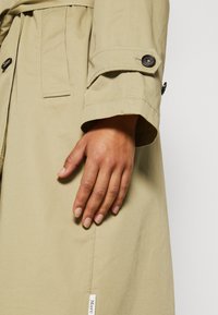 Main droite d'une personne portant un trench-coat beige avec des détails de brides boutonnées sur la manche et une étiquette de marque visible près de l'ourlet.