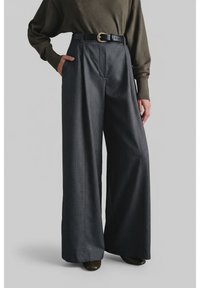 Kobieta ubrana w szerokie, ciemnoszare spodnie z czarnym paskiem, oliwkowozielony sweter i ciemne buty na tle jasnoszarego tła.