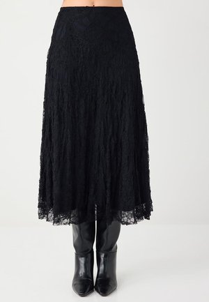 Zwarte kanten midi-rok met gegolfde zoom, gecombineerd met glanzende zwarte laarzen tot halverwege de kuit, tegen een effen lichte achtergrond.