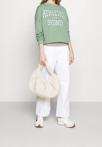 Felpa verde con stampa "ATHLETICS SYDNEY 1987", pantaloni bianchi a gamba larga e borsa cremosa e soffice. Le sneakers sono bianche con accenti chiari.