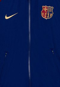 Nike zip-up jacka i blått tyg med guldlogga och FC Barcelona-emblem. Har två sidofickor med dragkedja.