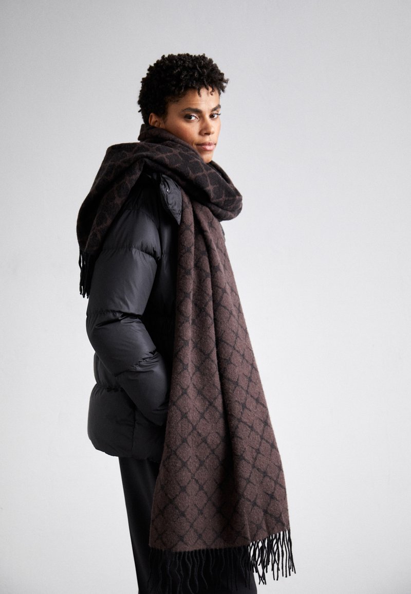 Brown patterned scarf made of soft fabric, draped around the neck. Features a geometric design and frayed edges, paired with a black puffer jacket.