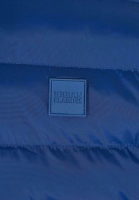 Mörkblå puffjacka med sydda horisontella paneler, försedd med en fyrkantig gummietsning som lyder "URBAN CLASSICS" med fet stil.