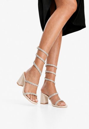 Piernas con sandalias beige de tacón ancho con tiras alrededor del tobillo adornadas con piedras brillantes, combinadas con un vestido negro.