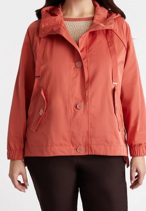 Kvinde iført en koralfarvet jakke med knapper, hætte, snørre, klaplommer, elastiske manchetter og sorte bukser.