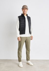 Man som bär beige keps, svart vadderad väst, långärmad vit skjorta, olivgröna byxor och vita sneakers som står mot en enkel bakgrund.