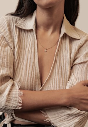 Frau trägt eine beige strukturierte Bluse mit hochgekrempelten Ärmeln und eine zarte goldene Halskette mit kleinem Anhänger. Arme verschränkt.