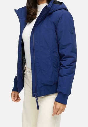 Vrouw draagt een blauw capuchonjack, open over een witte top en crème kleurige broek, staand met haar armen ontspannen langs haar zij.