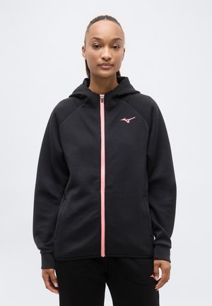 Naine, kes kannab musta kapuutsiga lukuga jope, millel on roosa lukk ja väikene roosa logo rinnal, seisab ühetaustal heleda tausta ees.