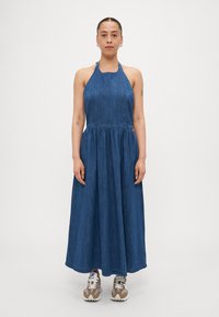 Denim halter-neck maxikjole med tætsiddende overdel og flagrende nederdel, med en samlet talje. Bæres med beige og hvide sneakers.