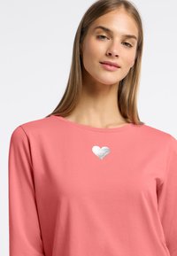 Różowa koszula z długim rękawem, wykonana z miękkiego materiału, z metalicznym srebrnym wzorem serca na klatce piersiowej. Prosty okrągły dekolt i luźny krój.