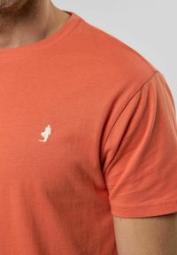 Krátký rukáv, tričko s kulatým výstřihem v korálově oranžové barvě z bavlny, s malým bílým logem na hrudi. Hladká textura a volný střih.