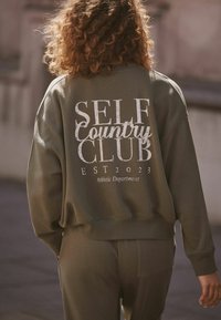 Alyvuogių spalvos džemperis su užrašu "SELF COUNTRY CLUB EST 2023" balta spalva nugaros pusėje, minkšta medžiaga, laisvas kirpimas, baigtos ribotos rankovės ir apatinė dalis.