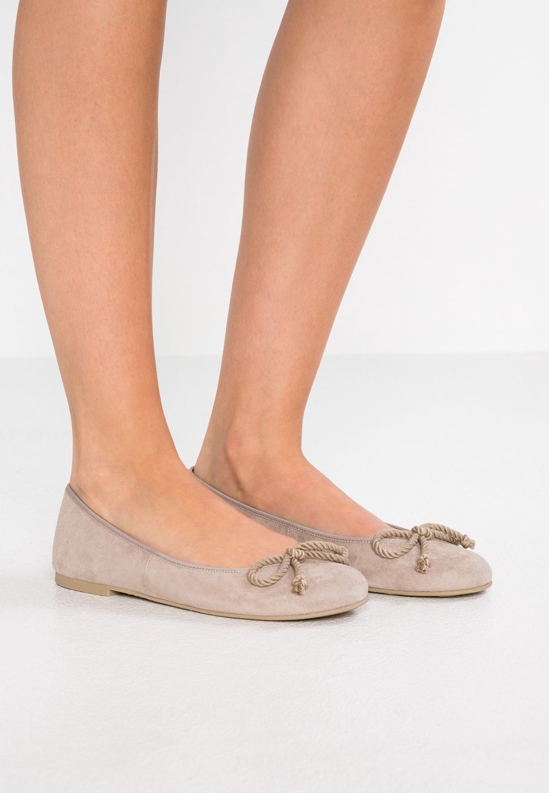 Beige ruskindsballetsko med rund tå, der har dekorative rebdetaljer foran og en flad, fleksibel sål.