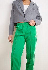 Blazer gris corto con un solo botón, sobre una camiseta sin mangas blanca, combinado con pantalones de deporte verdes que tienen franjas blancas y un logo de Adidas.