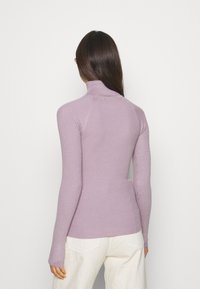 Kobieta o długich ciemnych włosach ubrana w dopasowany lawendowy sweter z prążkowanym golfem i białe spodnie, zwrócona tyłem na gładkim tle.