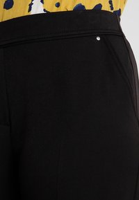 Pantalon noir à la texture lisse, coupe slim, avec un petit accent argenté sur la ceinture et des poches frontales inclinées.