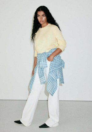 Femme aux longs cheveux bouclés portant un pull jaune, un pantalon blanc, des chaussures noires pointues et une chemise à carreaux bleue nouée autour de la taille.