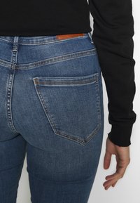 Jeans de mezclilla azul oscuro con un diseño ajustado, que presentan un parche de cuero en la cintura y dos bolsillos traseros con costura en contraste.
