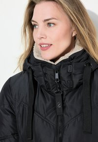 Giacca nera imbottita con colletto testurizzato color crema, dotata di collo alto, lacci e zip frontale con dettagli hardware a contrasto.