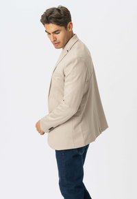 Beige blazer med slankt design, to frontlommer, spidse klapper og enkeltknaplukning, parret med mørke jeans.