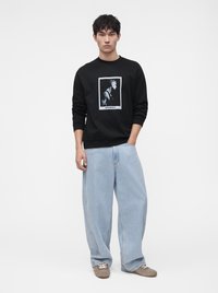 Homme portant un sweat-shirt noir avec un motif en noir et blanc, un jean ample bleu clair et des chaussures grises, debout avec une main dans la poche.
