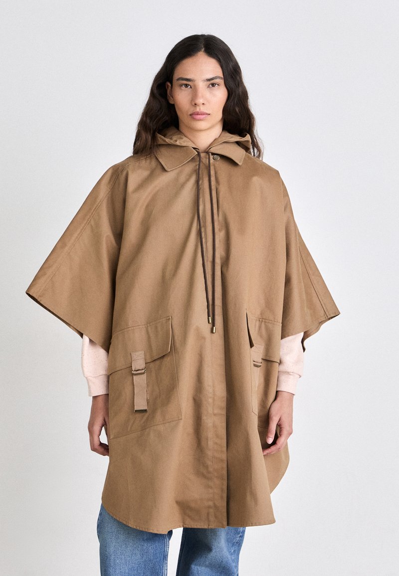 Poncho marron imperméable avec capuche, grandes poches avant, fermeture à cordon de serrage et coupe décontractée. Fabriqué en tissu coton texturé.