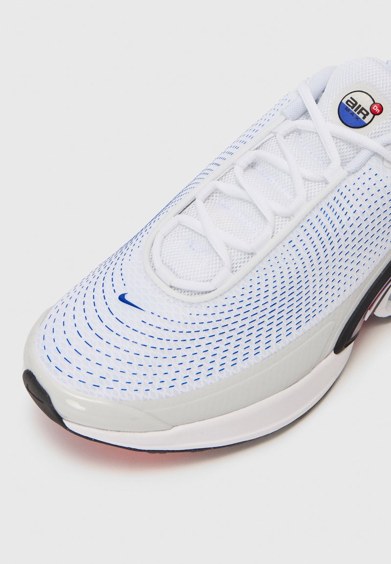 Zapatilla deportiva blanca con acentos azules, que cuenta con una parte superior de malla, un logo circular en la lengua, suela texturizada y perforaciones visibles para transpirabilidad.