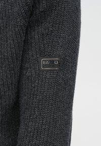 Pulover tricotat gri închis cu finish texturat și o etichetă dreptunghiulară neagră "Barbour International" pe mânecă.