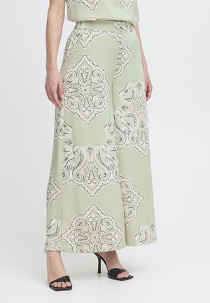 Femme portant un pantalon large vert clair avec de grands motifs floraux blancs et beiges et des sandales à talons noires.