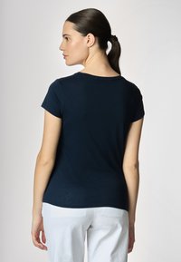 La donna, vista di spalle, indossa una maglietta blu navy e pantaloni bianchi, con i capelli raccolti. Uno sfondo neutro completa l'outfit.