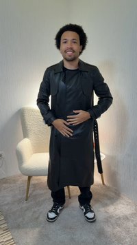 Muškarac s kovrčavom kosom, obučen u dugi crni kožni kaput i tenisice, stoji nasmiješen u sobi s tepihom, a iza njega se nalazi bijela fotelja.